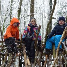 3 Kinder sitzen auf einen selbstgeebauten Unterstand aus Ästen. Im Hintergrund sind Bäume. Es liegt Schnee. 